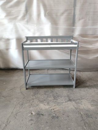 The Child Craft Flat Top Baby Changing Table in Cool Gray features two lower nursery storage shelves and a white changing pad, set on a concrete floor against a wall covered with plastic sheeting.