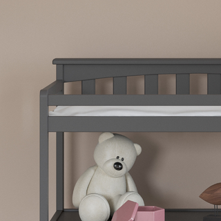 A large white teddy bear sits on the bottom bunk of a gray bunk bed, next to a pink toy and brown object, with the Child Craft Flat Top Baby Changing Table in cool gray nearby, all set against a beige wall.