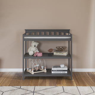 The Child Craft Flat Top Baby Changing Table in Cool Gray features baby-safe finishes, a white changing pad, and nursery storage holding a teddy bear, pink toy, books, decorative bowl, and wooden bead maze against a beige wall on a wood floor.