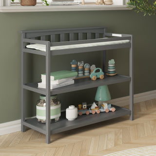 The Child Craft Flat Top Baby Changing Table in Cool Gray features baby-safe finishes and three shelves, holding a green changing pad, books, toys, and decor in a nursery with green walls and wood herringbone floors.