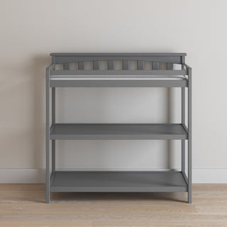 The Child Craft Flat Top Baby Changing Table in Cool Gray features baby-safe finishes, two storage shelves, and a padded changing area, shown against a white wall on a light wood floor.