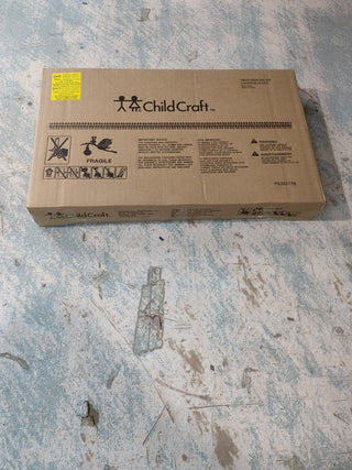 A closed cardboard box labeled "Child Craft" sits on a worn, light blue and white floor. The box, with handling symbols, warnings, and a yellow sticker in the top left, likely contains an Arch Top Baby Changing Table in Jamocha.