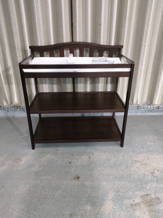 The Child Craft Arch Top Baby Changing Table in Jamocha features a dark wood finish, two lower shelves, and a white changing pad, complementing your nursery decor with beige curtains and a light-colored floor.