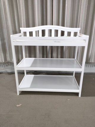 The Child Craft Arch Top Baby Changing Table in matte white features two open shelves for storage, standing on a beige carpet before light curtains—an ideal piece of nursery furniture.