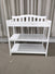 The Child Craft Arch Top Baby Changing Table in matte white, featuring a curved back and two open storage shelves, is shown as nursery furniture on a beige carpet in front of floor-length curtains.