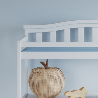 The Child Craft Arch Top Baby Changing Table in matte white stands against a pale blue wall, complementing nursery decor. A wicker apple-shaped basket and a wooden toy rest beside and under the table.