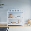 The Child Craft Arch Top Baby Changing Table in matte white, with a blue pad, wicker basket, wooden toys, books, and a stacking toy, sits against a light blue wall, paired with a striped rug, plant, and charming nursery decor.