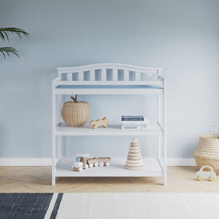 The Child Craft Arch Top Baby Changing Table in matte white, with a blue pad, wicker basket, wooden toys, books, and a stacking toy, sits against a light blue wall, paired with a striped rug, plant, and charming nursery decor.