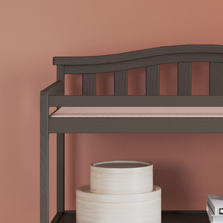 The Child Craft Arch Top Baby Changing Table in Dapper Gray stands against a peach-colored wall, perfect for a nursery. Two round, light-colored nested storage boxes sit partially under the table.
