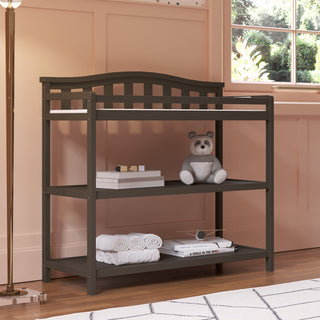 The Child Craft Arch Top Baby Changing Table in Dapper Gray features storage shelves holding a stuffed bear, books, folded towels, and a box. Curved railings frame the table, which sits near a window against pink paneled nursery walls.