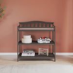 The Child Craft Arch Top Baby Changing Table in Dapper Gray features two storage shelves holding books, a wooden box, a toy abacus, and a red toy car, set against a terracotta wall with a leafy plant—ideal nursery furniture.