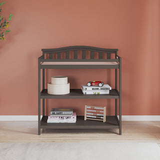 The Child Craft Arch Top Baby Changing Table in Dapper Gray features two storage shelves holding books, a wooden box, a toy abacus, and a red toy car, set against a terracotta wall with a leafy plant—ideal nursery furniture.