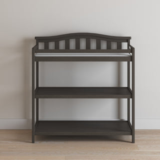 The Child Craft Arch Top Baby Changing Table in Dapper Gray features three open shelves, a slatted backrest, and raised sides for safety, offering stylish and functional nursery storage against a light wall and hardwood floor.