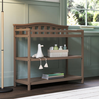 The Child Craft Arch Top Baby Changing Table in Dusty Heather includes two shelves for storing baby care items, bottles, and a basket. A plush toy sits on the middle shelf. The table is placed by a window with green paneling on the wall.
