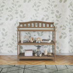 The Child Craft Arch Top Baby Changing Table in Dusty Heather stands against leafy wallpaper in a nursery with a patterned rug and parquet flooring, featuring two shelves for baby books, toys, a stuffed zebra, and a wooden abacus.