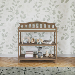 The Child Craft Arch Top Baby Changing Table in Dusty Heather stands against leafy wallpaper in a nursery with a patterned rug and parquet flooring, featuring two shelves for baby books, toys, a stuffed zebra, and a wooden abacus.