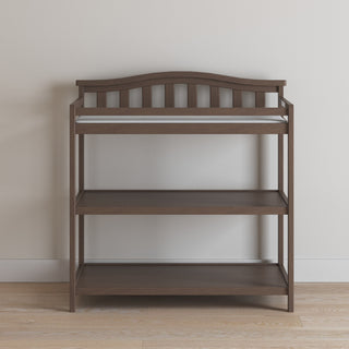 The Child Craft Arch Top Baby Changing Table in Dusty Heather, with a curved top rail and two open shelves for storage, stands against a light-colored wall on a wooden floor.