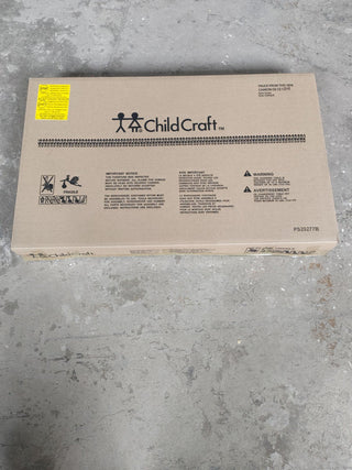 A sealed rectangular cardboard box labeled Child Craft sits on a concrete floor. It contains an Arch Top Baby Changing Table in Cool Gray, and displays several warnings, instructions, and symbols in black print.