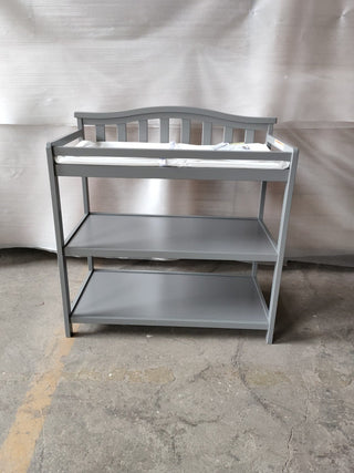 The Child Craft Arch Top Baby Changing Table in Cool Gray features a slatted back, two open shelves, and a padded changing mat, shown on a concrete floor against a neutral fabric backdrop.
