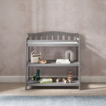 The Child Craft Arch Top Baby Changing Table in Cool Gray features three shelves, perfect for storing baby toys, books, and décor. It sits on a wood floor with a blue rug in front, set against a light beige textured wall.