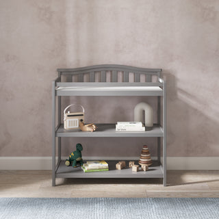 The Child Craft Arch Top Baby Changing Table in Cool Gray features three shelves, perfect for storing baby toys, books, and décor. It sits on a wood floor with a blue rug in front, set against a light beige textured wall.