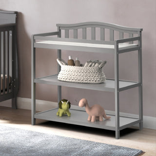 The Child Craft Arch Top Baby Changing Table in Cool Gray features three shelves, with a woven basket of baby supplies on the middle shelf and green lion and pink dinosaur toys on the bottom. A crib is partially visible to the left.