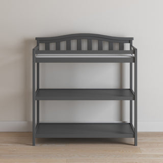 The Child Craft Arch Top Baby Changing Table in Cool Gray features a curved top rail, slatted sides, two open shelves, and a changing pad. It's shown against a white wall on a wooden floor.