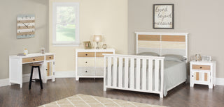 A bedroom featuring a white wooden bed, matching nightstand and dresser, plus the Child Craft Rockport Desk in Sandstone. Decor includes a lamp, clock, books, framed art, a "loved beyond measure" sign, hardwood floor, and beige rug.