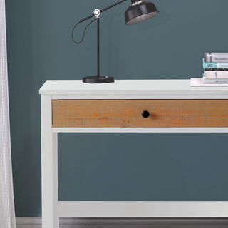 The Child Craft Rockport Desk in Sandstone features a single wooden drawer, paired with a black desk lamp and a small stack of books, all set against a blue wall with white curtains partially visible on the left.