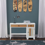 The Child Craft Rockport Desk in Sandstone, featuring a drawer and cabinet, stands against a teal wall with books and a black lamp on top. Four surfboards hang above; white curtains surround, and a blue patterned rug lies below.