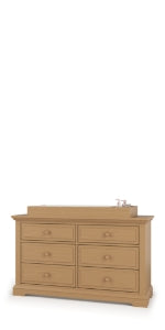 The Child Craft Legacy Changing Table Topper in Biscotti sits atop a light brown wooden dresser with six drawers and round handles, featuring a raised tray and small container set at the back right corner against a plain white background.