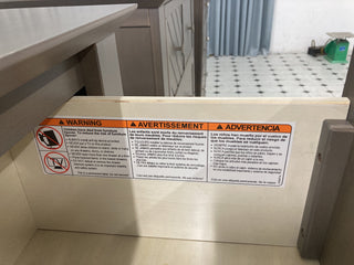 Inside the Atwood 4-in-1 Convertible Crib by Child Craft, a warning label in English, French, and Spanish advises to keep children away to prevent tipping hazards. Includes a graphic of a child with a crossed-out tip-over symbol.
