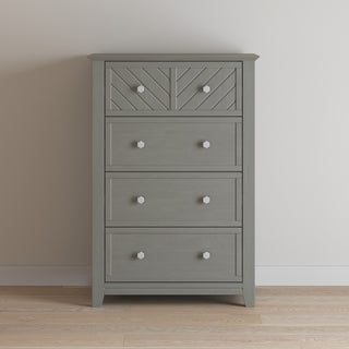 The Child Craft Atwood 4-Drawer Chest in Lunar Gray features white hexagonal knobs, a patterned top drawer, and three plain drawers. Shown on a light wood floor against an off-white wall.