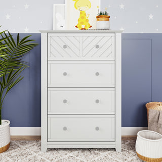 The Child Craft Atwood 4-Drawer Chest in matte white stands against a blue and white star-patterned wall, adorned with decorative items like a giraffe print, a plant, and a basket.