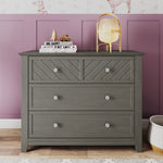 The Child Craft Atwood 3-Drawer Dresser in Lunar Gray stands against a mauve paneled wall, topped with a lamp, books, and figurines; a basket is partially visible on the floor to the right.