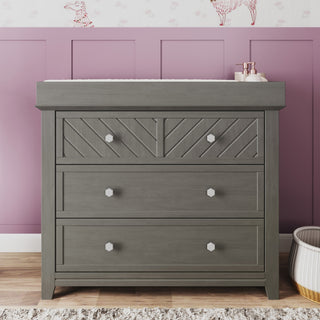 The Child Craft Atwood 3-Drawer Dresser in Lunar Gray stands against a mauve paneled wall with animal-print wallpaper, topped with a small tray and bottle, while a woven basket sits on the floor beside it.