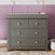 The Child Craft Atwood 3-Drawer Dresser in Lunar Gray stands against a mauve paneled wall with animal-print wallpaper, topped with a small tray and bottle, while a woven basket sits on the floor beside it.