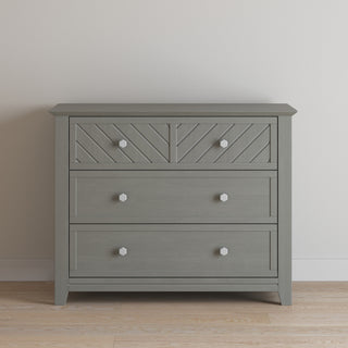 The Child Craft Atwood 3-Drawer Dresser in Lunar Gray features geometric patterns on the top drawer and white hexagonal knobs. It stands against a plain white wall on a light wood floor.