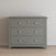 The Child Craft Atwood 3-Drawer Dresser in Lunar Gray features geometric patterns on the top drawer and white hexagonal knobs. It stands against a plain white wall on a light wood floor.