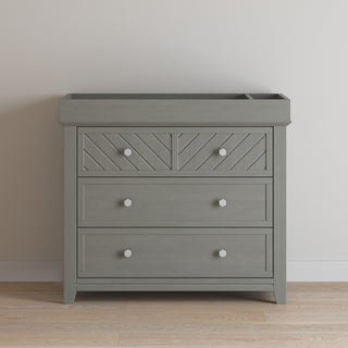 The Child Craft Atwood 3-Drawer Dresser in Lunar Gray features geometric patterns on the top drawer and white knob handles, standing against a plain white wall on a light wood floor.