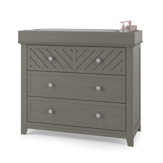 The Child Craft Atwood 3-Drawer Dresser in Lunar Gray features a chevron-patterned top drawer, a changing pad on top, and two bottles on the right side, perfect for storing lotion or soap.