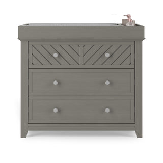 The Child Craft Atwood 3-Drawer Dresser in Lunar Gray features a textured front, three drawers, a cushioned changing pad on top, and a pink soap dispenser set at the back right corner.