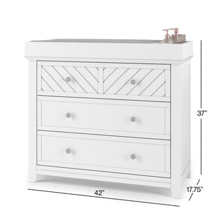 White dresser with three drawers, a geometric top drawer, and decorative knobs. Pair with Child Craft's F01792 Universal Changing Table Topper in Matte White to convert it into a baby changing station. 42"W x 17.75"D x 37"H. Two bottles on top right corner.