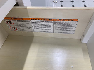A warning label in English, French, and Spanish is inside the Atwood 3-Drawer Dresser by Child Craft, advising users to secure the furniture to the wall to prevent tip-over accidents. The label features safety icons and bold warning text.