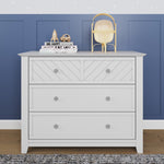 The Child Craft Atwood 3-Drawer Dresser in matte white stands against a blue paneled wall. On top are stacked books, a gold lamp, a rocket ornament, and a brown alien figurine. The room features a wooden floor.