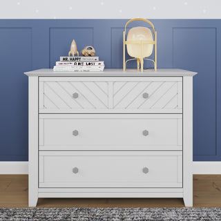 The Child Craft Atwood 3-Drawer Dresser in matte white stands against a blue paneled wall. On top are stacked books, a gold lamp, a rocket ornament, and a brown alien figurine. The room features a wooden floor.