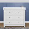 The Child Craft Atwood 3-Drawer Dresser in matte white stands by a blue paneled wall, with a gray rug in front and a pink soap dispenser on top.
