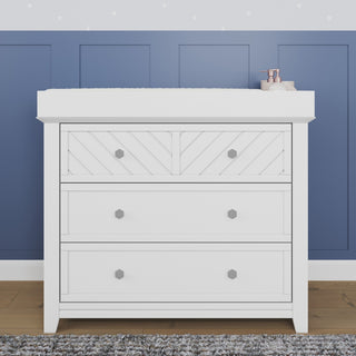 The Child Craft Atwood 3-Drawer Dresser in matte white stands by a blue paneled wall, with a gray rug in front and a pink soap dispenser on top.