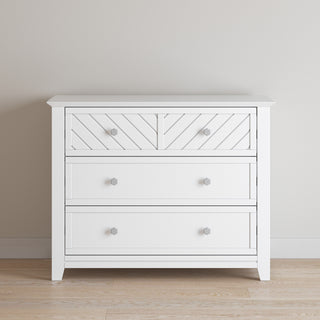 The Child Craft Atwood 3-Drawer Dresser in matte white features geometric detailing on the top drawer and round knobs, shown standing on a wooden floor against a light-colored wall.