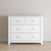 The Child Craft Atwood 3-Drawer Dresser in matte white features geometric detailing on the top drawer and round knobs, shown standing on a wooden floor against a light-colored wall.